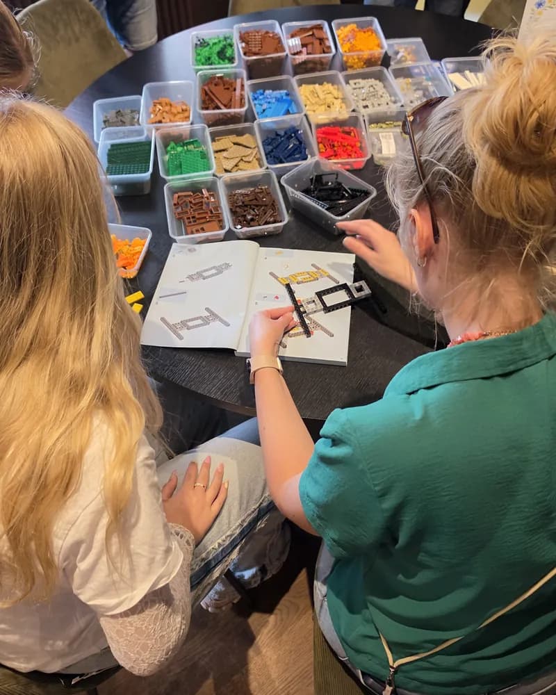 Vänner bygger tillsammans vid ett bord med LEGO®-bitar sorterade i lådor och en bygginstruktion